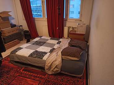 The image depicts a small room with a bed, red curtains, and a window, featuring a patchwork quilt and a red patterned rug on the floor.