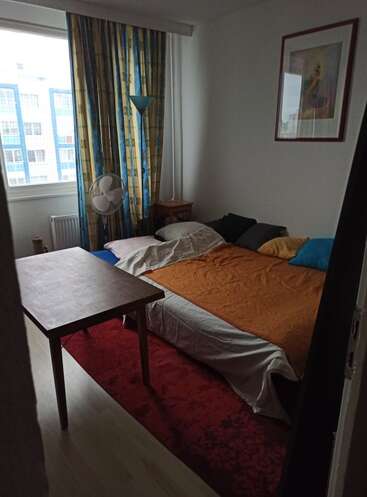 The image depicts a small bedroom with a bed, table, and window, featuring a vibrant red rug and a mix of colorful curtains and bedding.
