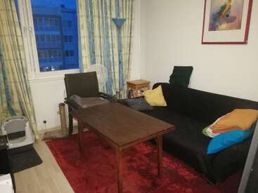 The image depicts a cozy living room with a black couch, wooden coffee table, and red rug, featuring a window with curtains and a cityscape outside.