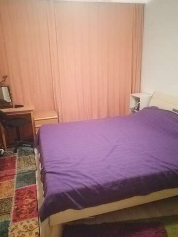 The image depicts a bedroom with a bed, desk, and chair, featuring a purple bedspread, pink curtains, and a colorful rug, set against a white wall.