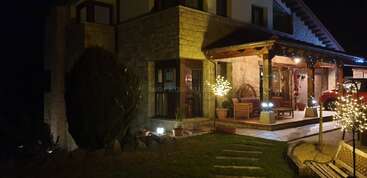 This image shows a cozy stone house at night, warmly lit with string lights, decorated porch, illuminated trees, potted plants, and a red car parked nearby.