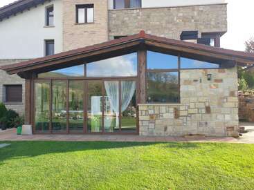 This image shows a modern house extension with large glass windows and doors, a sloped roof, stone walls, green lawn, and scenic reflections of nature.