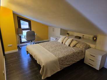 This cozy attic bedroom features a sloped ceiling, yellow accent wall, a double bed with patterned bedding, desk with chair, wooden floor, and minimalist decor.