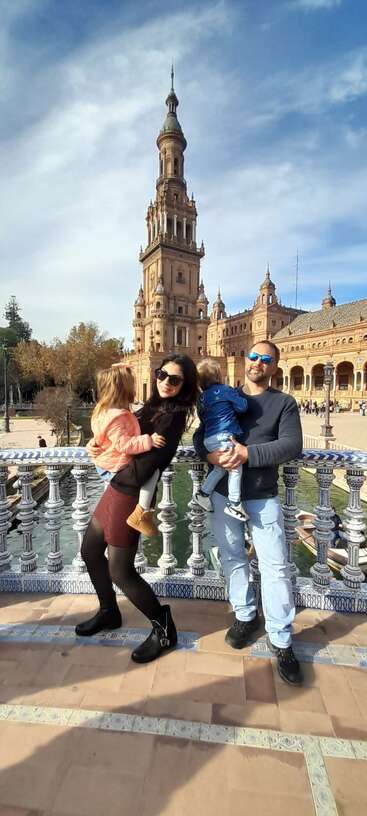 A family of four poses happily on a decorative bridge at Plaza de España in Seville, Spain, with the iconic tower and blue sky behind them.