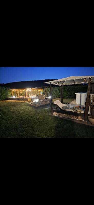 A cozy outdoor setting at dusk features a lit gazebo with people relaxing, a canopy bed, lush green grass, and a pool, surrounded by tall hedges.