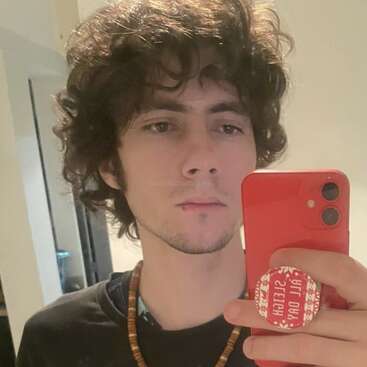 A young person with curly brown hair takes a mirror selfie using a red phone. They're wearing a necklace and a black shirt, standing in a simple room.