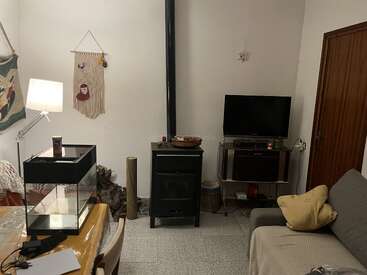 A cozy living room with a wood stove, television, sofa, lamp, aquarium on a table, wall hangings, and a wooden door. Relaxed atmosphere and decor.