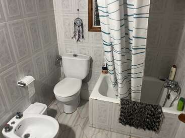 This is a small bathroom with a toilet, bidet, bathtub, striped shower curtain, window, dreamcatcher, toiletries, and a textured floor mat hanging on the tub.