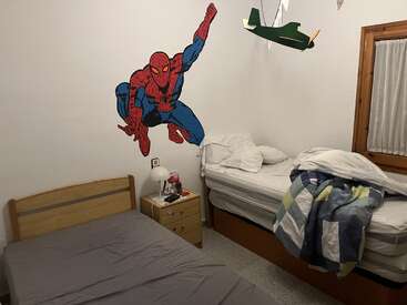 This room features two unmade beds, a nightstand with a lamp, a large Spider-Man wall decal, and a hanging airplane model above one bed. Cozy atmosphere.