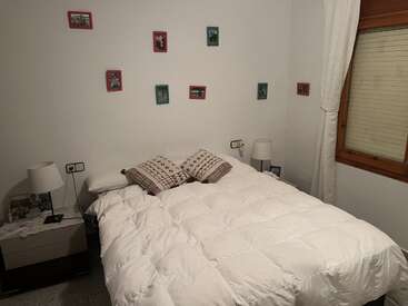 A cozy bedroom features a neatly made bed with two pillows, nightstands with lamps, framed photos on the wall, and a window with white curtains.