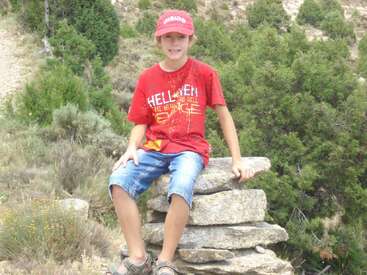 Ein Junge mit roter Mütze, rotem T-Shirt und Jeansshorts sitzt draußen auf einem Steinhaufen, umgeben von grünen Büschen und unwegsamem Gelände.