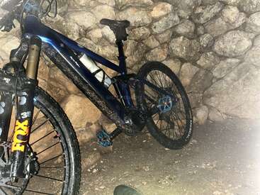 Ein blau-schwarzes Mountainbike lehnt an einer rauen Steinmauer. Das Fahrrad ist schmutzig, was darauf hindeutet, dass es kürzlich auf schlammigem Gelände benutzt wurde, und hat eine FOX-Federung.