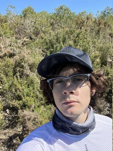 Ein junger Mensch mit Brille, schwarzer Mütze und Halswärmer macht draußen ein Selfie. Im Hintergrund sieht man grüne Büsche und Bäume unter einem strahlend blauen Himmel.