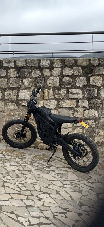 Ein schickes schwarzes E-Bike steht auf Kopfsteinpflaster und lehnt an einer Steinmauer mit einem Metallgeländer darüber. Der Himmel sieht bewölkt aus.