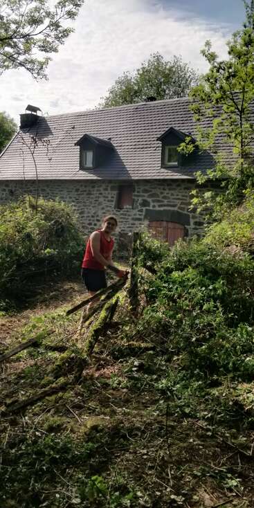 Une personne vêtue d'un débardeur rouge est en train de débroussailler devant une maison en pierre au toit de tuiles grises, entourée de verdure et d'arbres.