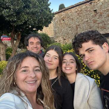 A group of five people smile for a selfie outdoors, standing close together. Behind them are yellow flowers, a tree, and an old stone building.