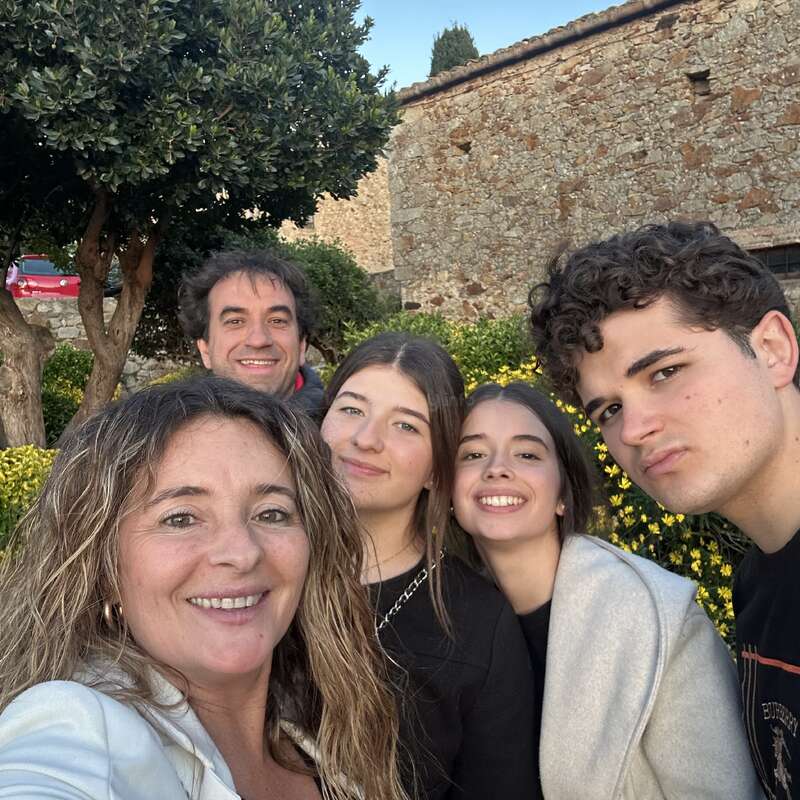 A group of five people smile for a selfie outdoors, standing close together. Behind them are yellow flowers, a tree, and an old stone building.
