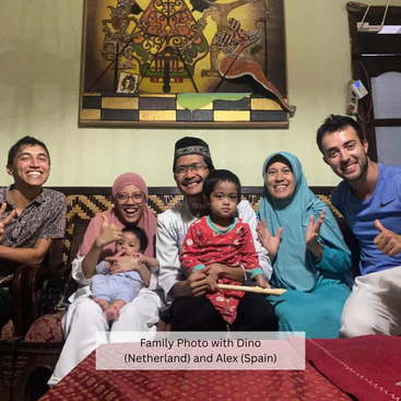 Ein fröhliches Familienfoto mit sieben Personen, darunter zwei Kinder. Alle lächeln, sitzen auf einer Couch und zeigen Wärme und Zusammengehörigkeit unter traditioneller Wandkunst.