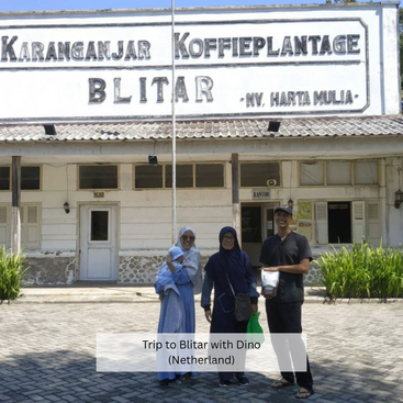 Tres personas posan delante del histórico edificio Karanganjar Koffieplantage en Blitar, Indonesia. El tiempo es soleado, lo que resalta sus expresiones alegres.