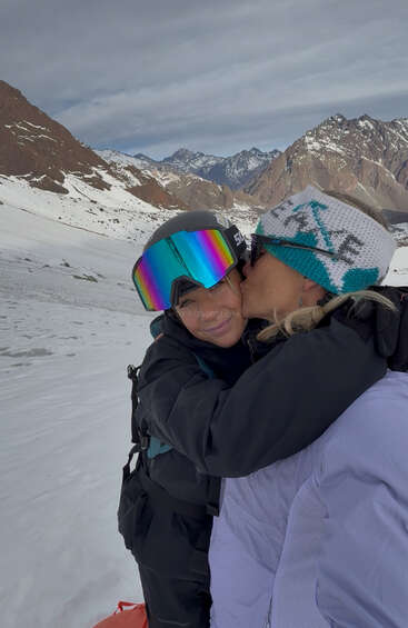 Two people embrace on a snowy mountain. One kisses the other’s cheek, both wearing winter gear and goggles, with beautiful snow-covered peaks in the background.
