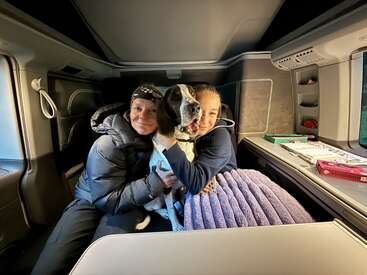 Two women sit together inside a cozy camper van, hugging a black and white dog, with a purple blanket and some books on the table nearby.