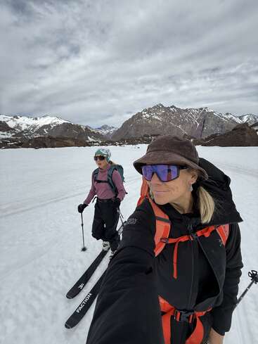 Two women are skiing on a snowy landscape, surrounded by rugged mountains. Both wear winter gear, backpacks, and sunglasses. The sky is mostly cloudy, creating a dramatic scene.