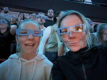A group of people wearing blue 3D glasses, with two women in the foreground smiling, enjoying an event in a crowded indoor venue with excitement.