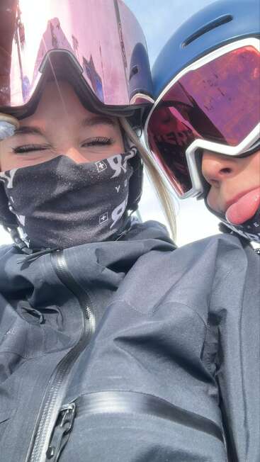 Two friends in ski gear, wearing helmets, goggles, and face coverings, take a close-up selfie outdoors. One sticks their tongue out playfully, snow visible reflected in goggles.