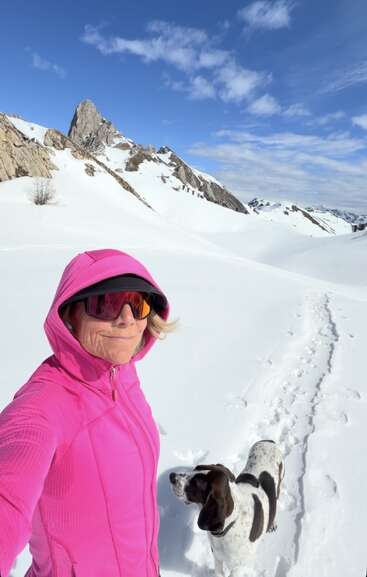 A woman in a bright pink jacket and sunglasses stands in snowy mountains, accompanied by a black-and-white dog under a bright blue, lightly clouded sky.