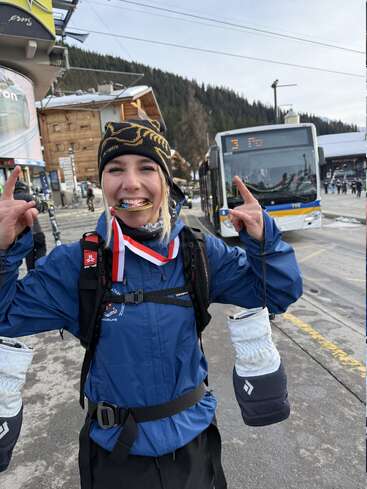 A smiling woman in winter gear stands outdoors, holding up her hands. She bites a colorful medal with a ribbon, celebrating, with buses and mountains in the background.