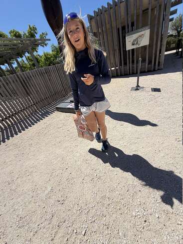 A young woman wearing sunglasses, a navy jacket, and shorts walks outdoors on a sunny day, holding a colorful bag. Wooden structures and signage are behind her.