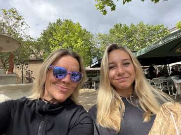 Deux femmes aux cheveux blonds sourient pour un selfie en plein air. L'une d'elles porte des lunettes de soleil violettes. Derrière elles, on aperçoit des arbres verts, une fontaine et un café avec des parasols.
