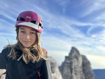 The image depicts a young woman wearing a pink helmet, standing in front of a mountainous landscape under a blue sky with wispy clouds.