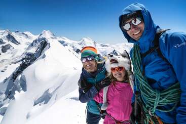 The image depicts three individuals standing together in a snowy mountainous landscape, all wearing winter attire and smiling at the camera.