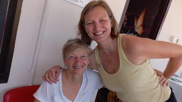 Duas mulheres estão sorrindo juntas dentro de casa. Uma está sentada, vestindo uma blusa branca, enquanto a outra está em pé ao lado dela, vestindo uma regata bege. O ambiente é acolhedor.