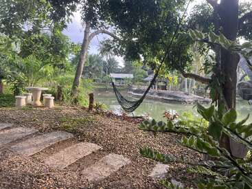 Uma cena tranquila no jardim mostra uma rede entre as árvores, uma mesa de pedra com bancos, um lago com vegetação exuberante e um pequeno pavilhão do outro lado da água.