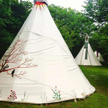 La imagen muestra dos tipis blancos con dibujos pintados, situados en una zona de hierba rodeada de árboles, que evocan un ambiente sereno y tranquilo.