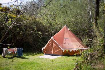 La imagen muestra una gran carpa con estructura de madera, mesa y sillas en su interior, sobre un fondo natural de árboles y una masa de agua. La tienda está situada en una zona de césped.