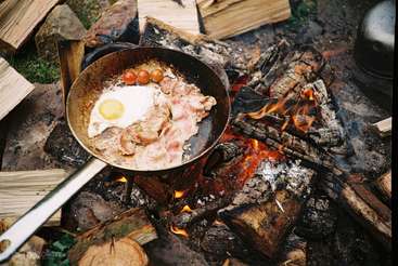 La imagen muestra una hoguera con una olla colgada sobre ella, rodeada por un grupo de personas sentadas sobre troncos, disfrutando de una comida juntos. La escena es tranquila e idílica.