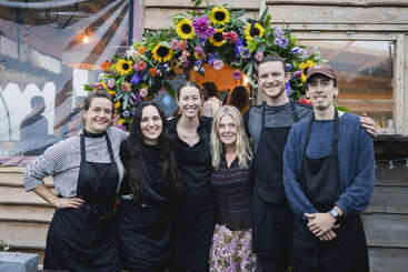 Six smiling people pose in front of a building adorned with a colorful floral arch. Most wear black aprons, suggesting they are a team or staff.