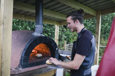 A man is smiling while preparing to place dough into a wood-fired brick oven outdoors, surrounded by greenery, likely making pizza or bread in a rustic setting.