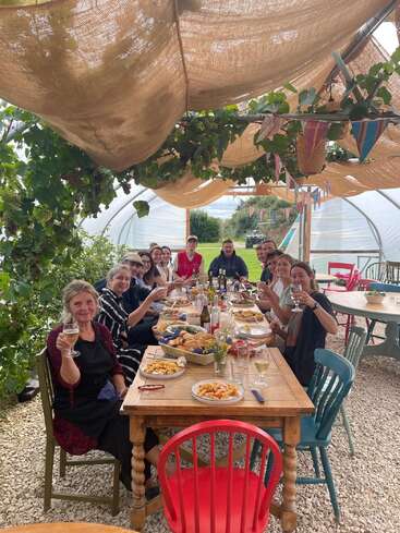 A group of people are gathered around a rustic outdoor table, enjoying food and drinks, smiling and raising glasses, under a leafy, beautifully decorated canopy.