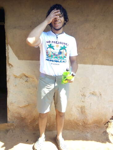 A man stands in front of a tan wall, wearing a white t-shirt with a colorful design, khaki shorts, and white Crocs, holding a green drink bottle.