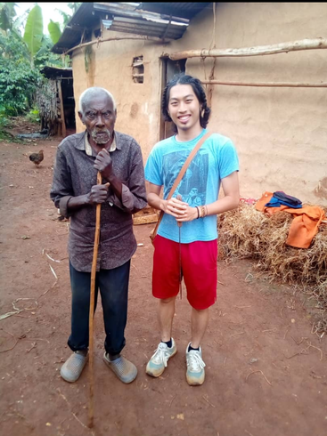 The image depicts a young man standing alongside an elderly gentleman in front of a rustic mud hut, conveying a sense of warmth and connection.