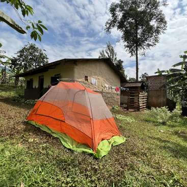 The image depicts a vibrant orange and green tent situated in front of a rustic house, surrounded by lush trees and a serene blue sky with white clouds.
