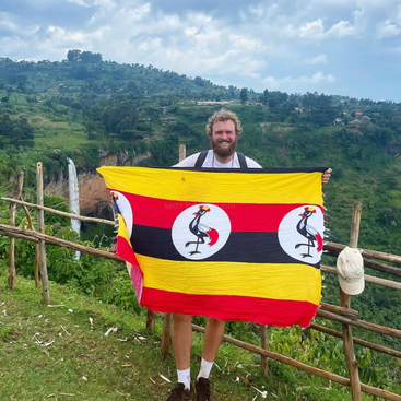 A man stands in front of a wooden fence, proudly holding a Uganda flag with a serene mountainous landscape in the background on a cloudy day.
