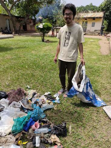 A man stands in a grassy area, holding a bag of trash, with a pile of litter on the ground in front of him, amidst a serene outdoor setting.