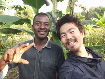 The image shows two men standing together in front of a banana tree, with the man on the left making a peace sign with his hand.