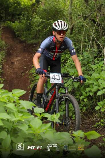 Ein Radfahrer mit Helm und Sonnenbrille fährt auf einem Mountainbike mit der Nummer „43“ über einen üppig grünen Waldweg – konzentriert und entschlossen während eines Wettkampfs.