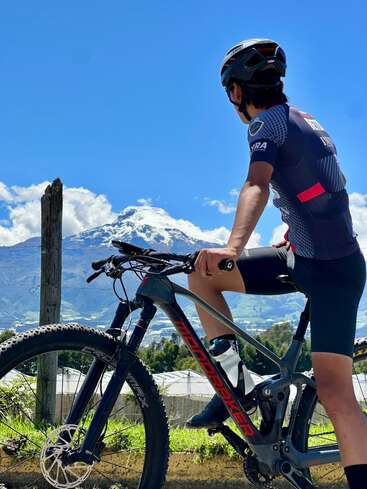 Ein Radfahrer mit Helm und Sportkleidung ruht sich auf seinem Mountainbike aus und genießt den malerischen Blick auf einen schneebedeckten Berg unter einem klaren, strahlend blauen Himmel.
